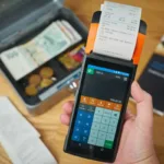 Tangan memegang mesin kasir yang mencetak nota transaksi dengan uang tunai dan koin di samping
